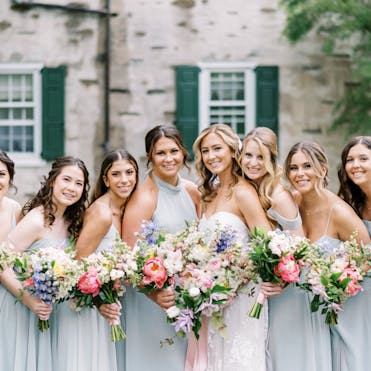 A bride with seven smiling bridesmaids, holding colorful floral bouquets outside the Appleford Estate in Villanova, PA