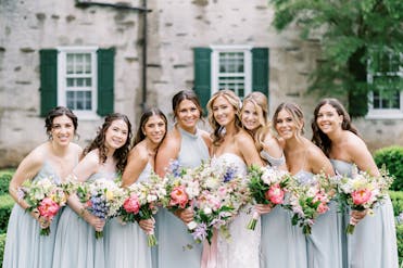 A bride with seven smiling bridesmaids, holding colorful floral bouquets outside the Appleford Estate in Villanova, PA