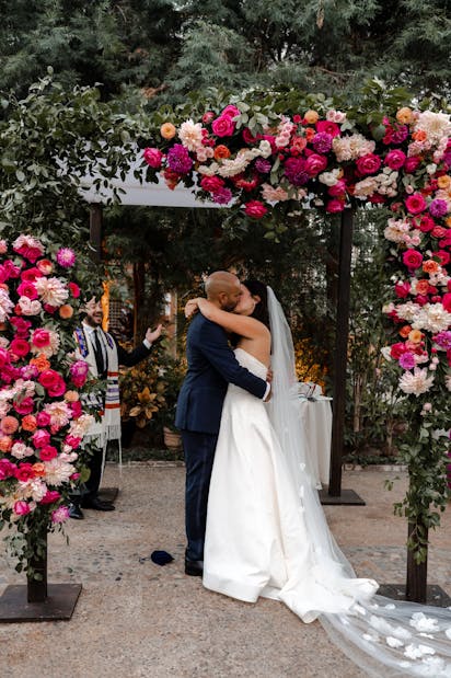 Under the cover of a pink floral arch, a pair of newlyweds share their first kiss