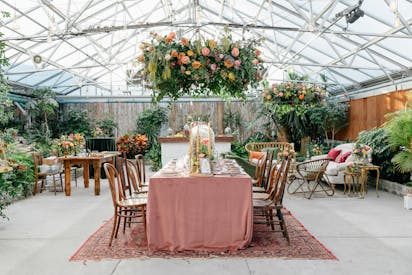 A quaint outdoor dinner table, surrounded by lush flowers and plants, is set for a fancy meal