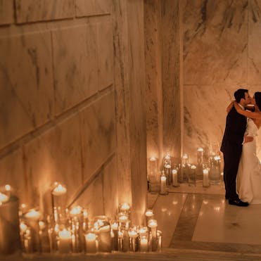 A freshly-married bride and groom embrace in a long marble hallway, illuminated by a line of candles