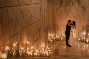 A freshly-married bride and groom embrace in a long marble hallway, illuminated by a line of candles